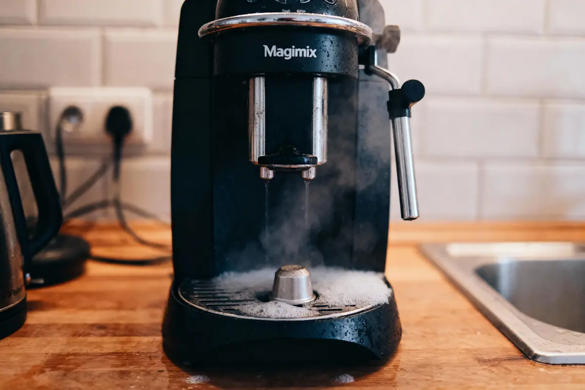 Machine à café sur un plan de travail, avec vapeur et débordement de mousse. Fond de cuisine carrelé.