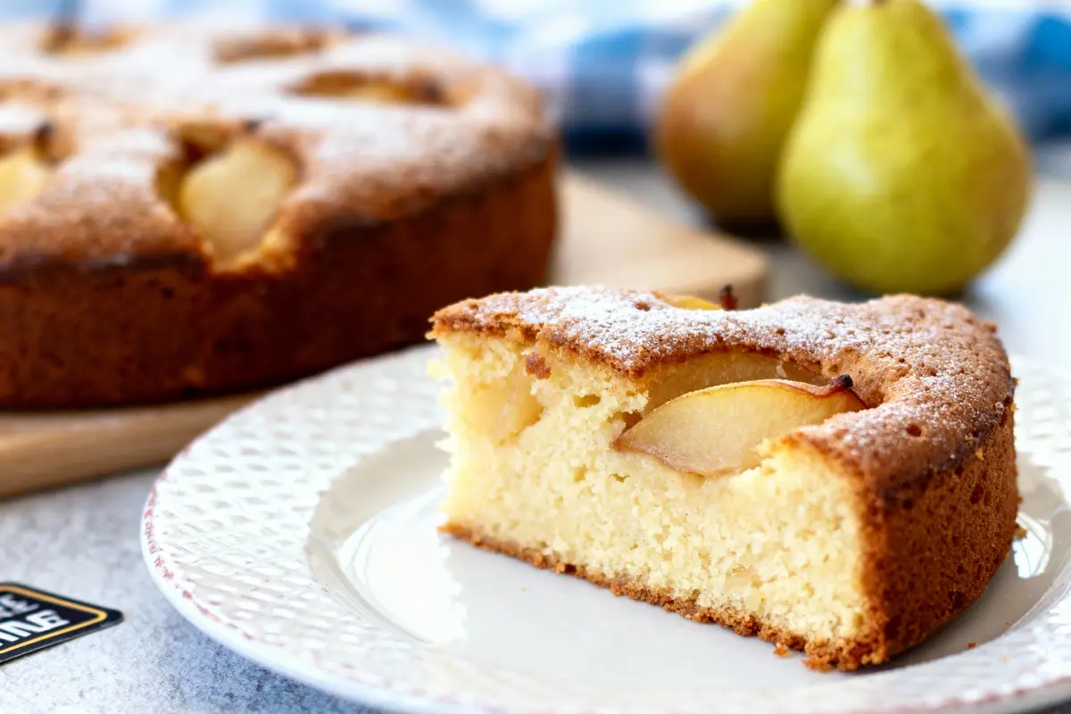 Part de gâteau poire sur assiette blanche, gâteau entier et poires floues en arrière-plan.