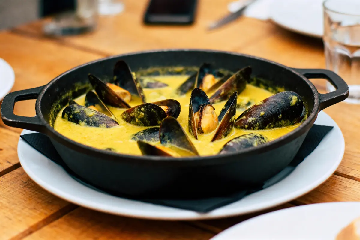 Plat de moules dans une sauce jaune crémeuse, servi dans une casserole noire sur table en bois.
