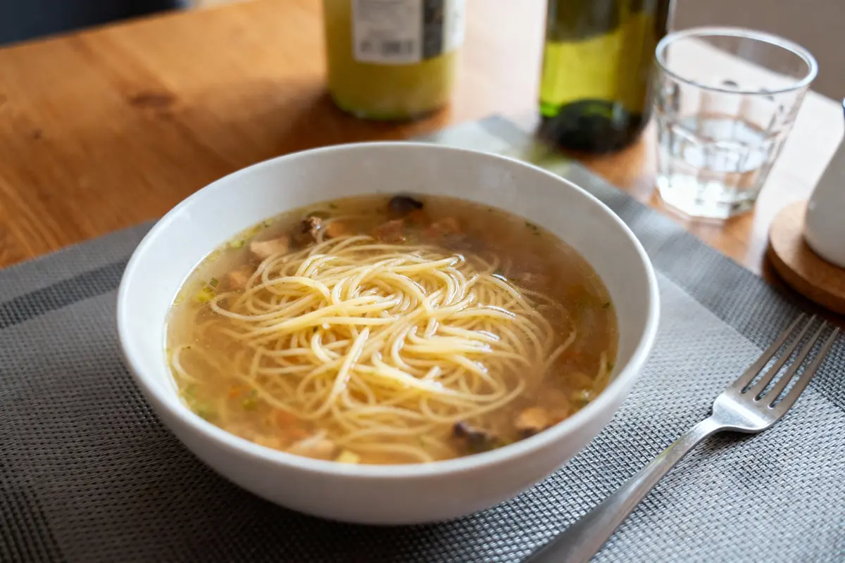 Bol de soupe avec nouilles, champignons, assiette grise, fourchette, bouteille et verre sur table en bois.