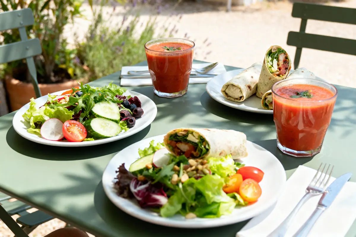 Salades et wraps avec gaspacho, servis sur une table extérieure verte, sous le soleil.