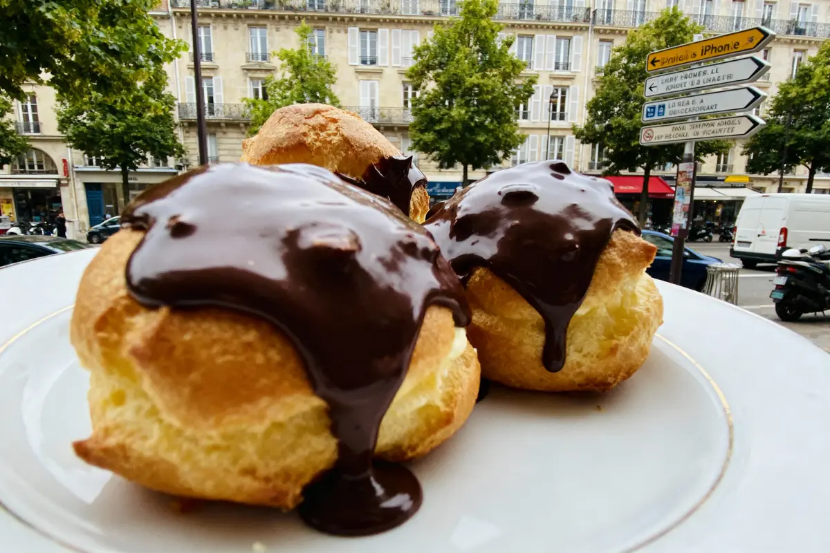 Choux à la crème chocolatés sur une assiette, arrière-plan de bâtiments parisiens et panneaux de signalisation.