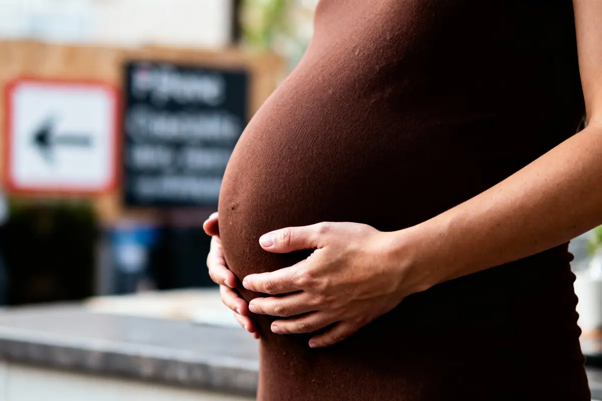 Ventre de femme enceinte en robe marron avec des mains posées dessus, arrière-plan flou urbain.