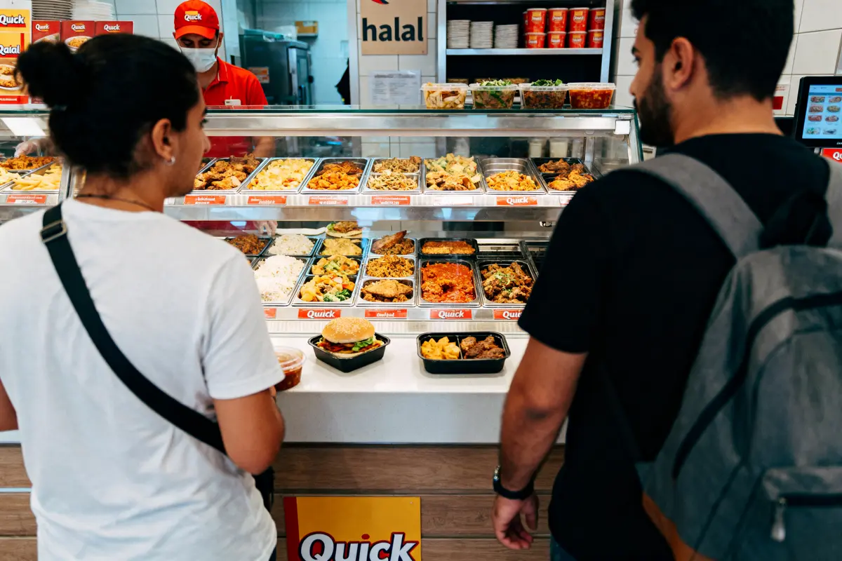 Deux personnes devant un comptoir de restauration rapide halal, plats variés exposés, personnel en arrière-plan.