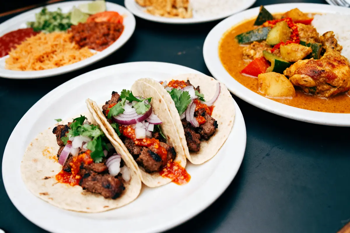 Trois tacos garnis de viande, oignons, coriandre et sauce rouge sur une assiette blanche, avec plats en arrière-plan.