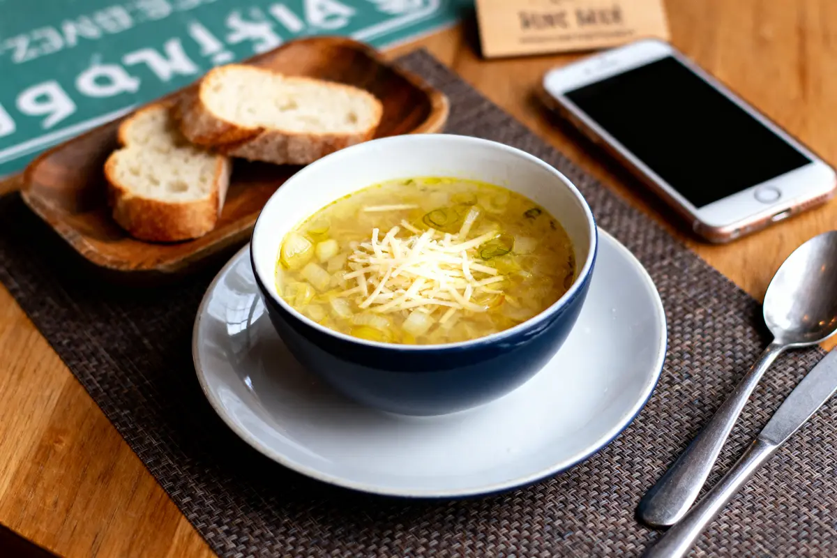 Bol de soupe avec fromage râpé, pain tranché, téléphone et couverts sur une table en bois.